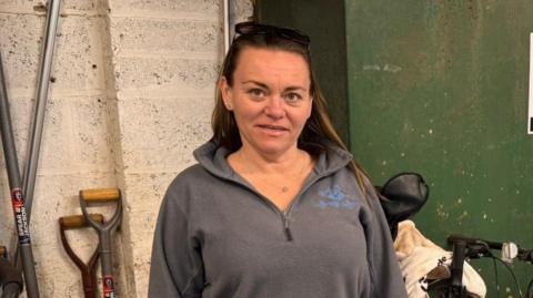 Rachel Douglas is a woman in her 40s wearing a grey top and has long brown hair. She is standing in front of a green tank - on the top is a guage which has an arrow pointing to empty 