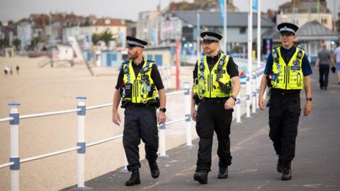 Three police officers walking along a beach area on a cloudy day. Some people behind them are walking away, others can be seen on the sand.