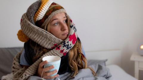 A woman sits in bed with scarves around her head and holding a cup of tea.