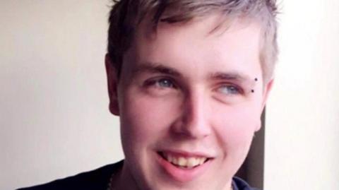 A close up picture of a young man, Darryl Thompson, who looks away to his left, smiling warmly. He has pale blue eyes, medium length textured light brown hair and a bar piercing in his left eyebrow. He wears a navy blue top and has a silver chain around his neck. 