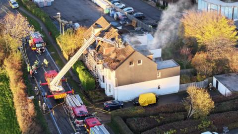 A drone shot of the fire coming from the house roof and fire engines in the road outside the houses in Brighton Road. One fire engine has a platform being held above the house