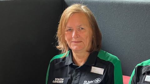 A picture of Viv Moore smiling looking at the camera wearing a StJohn's Ambulance black top with green shoulders, a Youth Leader badge.