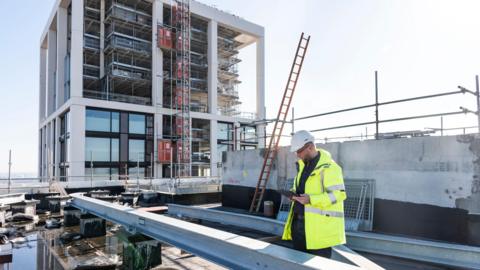 A man wearing a white hard hat and a yellow high viz jacket is standing on a building site. In front of him is a steel girder and to the left is a half-completed building with the floors exposed. In the background is a ladder and scaffolding.