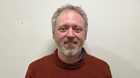 Tony Blake is standing in the middle of the shot wearing a rusty red jumper, with a blue shirt underneath. He has grey blonde hair, and a beard and moustache. 