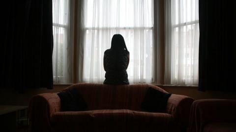 A woman sits on the back of a sofa, looking out of the window in fear of her partner, coming home after football. 