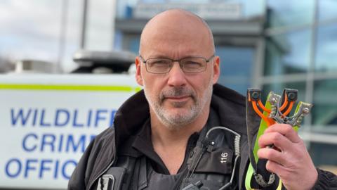 DC Aaron Flint, wearing a black police uniform stands in front of a police car holding two catapults. One catapult is orange and another is silver coloured.