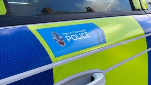 A close up shot of the outside door of a Devon & Cornwall Police car. The forces branding is on the outside in a blue rectangle. The regular police colours of yellow and blue are wrapped around the vehicle.