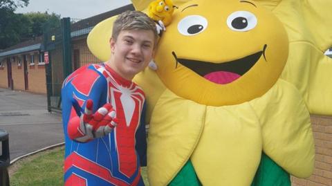 A teenage boy with short blonde hair is wearing a spiderman costume and standing next to a giant yellow cartoon flower 