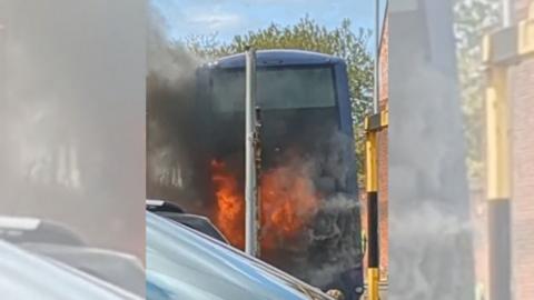 The back of a blue bus. The picture has been taken from behind the vehicle and flames can be seen coming from it. 