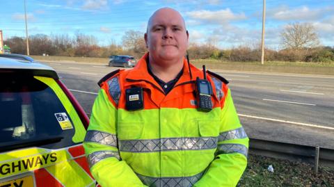 Traffic officer Robert Oates is standing by the M1. There are cars behind him on the motorway. He is wearing a fluorescent jacket with a radio and a bodycam attached to it.