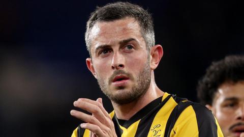 A photo of footballer Ben Garrity looking sternly into the distance while clapping on a pitch and wearing a yellow and black striped Port Vale Football Club shirt. A team-mate is blurrily visible over Garrity's left shoulder