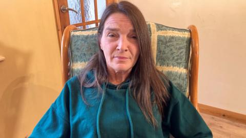 A woman sat down on a high backed chair looking straight ahead. She has long, dark brown hair and is wearing a teal coloured hooded jumper. 