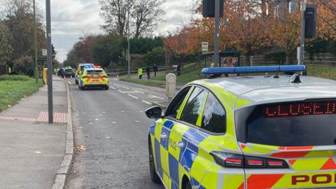 A police car blocks a road with the word "closed" shown on a display in the rear window. More police cars and officers are stood further down the road.
