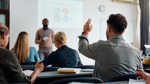 A student has his hand raised in a lecture. A teacher is standing at the front near a board with information on it. Several students are seen writing and looking at text books. 