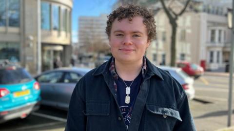 A young man with medium-length, brown, curly hair looks into the camera lens. He is wearing a navy jacket over and red and white checked shirt and blue t-shirt. He is wearing a necklace with two silver pendants.