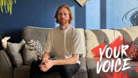 Male sitting on sofa wearing T shirt and dark trousers. The wall is blue and the sofa is grey. Light is coming in from the window on the right of the picture