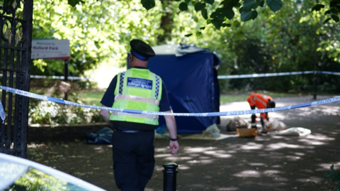 A blue and white cordon of police tape blocks off the entrance to a Manchester park. A uniformed Police Community Support Officer has his back to the camera, and in the background is a man in an orange hi-vis jacket standing beside a blue forensics tent, scooping matter into plastic bags. 