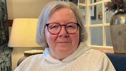 A woman with grey hair and glasses is wearing a white hoody and sat in an army chair near a lamp that is switched on
