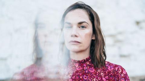 A woman stares at the camera with a sad look. She is wearing a pink floral dress. A reflection of her image is imposed to the left-hand side.