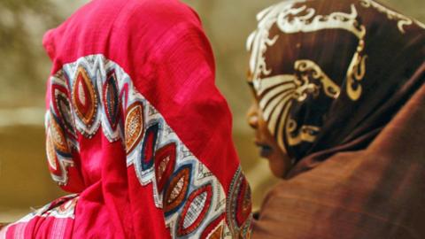 Two women in Zamzam camp in North Darfur seen from the back, wearing head scarves - one with a pink design and the other with a brown and cream design