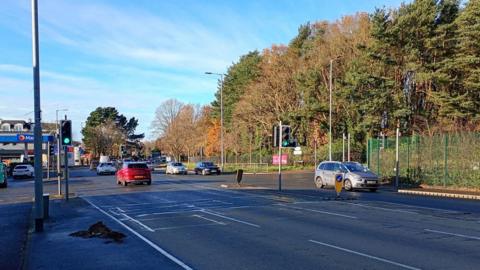 Various cars travelling on the Upper Newtownards Road in east Belfast on 30 November, 2025, near traffic lights and a Maxol service station with a blue and white sign in the distance. There are trees to the right and a green fence.