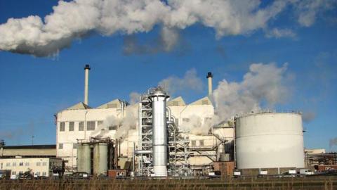 A factory site with a large metal cylinder and buildings can be seen from the other side of a river, with clouds of steam rising into the air on a sunny day