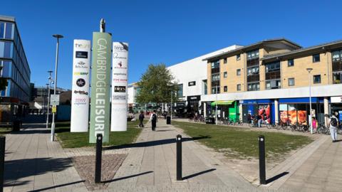 Cambridge Leisure Park, with a path and grassed area through the centre of it. To the left is the edge of a hotel. To the right are shops and a cinema. A big sign saying "Cambridge Leisure" in the foreground shows the different facilities based at the site. There are some people walking around and many bikes are parked near the shops.