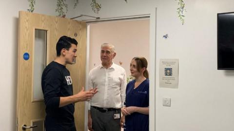 Sunderland footballer Luke O'Nien wearing a black jumper with a white log speaks to consultant Majd Abuarb who is wearing a white shirt and unit manager Emily Cameron who is wearing blue 