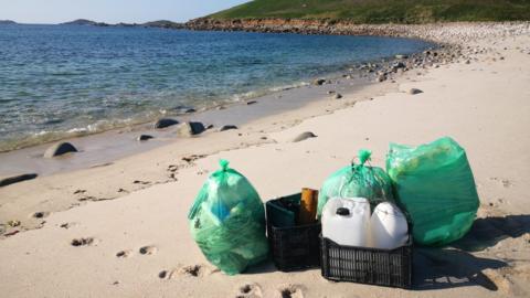 Bagged up marine debris on the sand of a beach. The sand is golden and there are footprints near the green bagged up items. The sea is behind it and is blue and clear. 