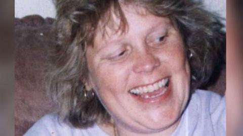 Debbie Griggs looks off camera and is smiling. She is wearing a pale coloured t-shirt and has short light brown hair.