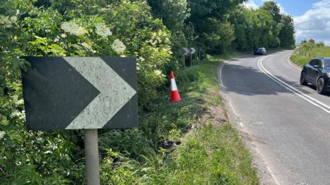 A black sign with an arrow on it on the side of the A36 in Wiltshire. The road next to the sign curves to the right. There are two cars travelling in opposite directions on the road. There is a traffic cone on the grass verge near the sign.
