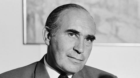 A black and white photo of Sir Alf Ramsey during a press conference. He has dark hair that is slicked back and he wears a suit, shirt and tie. A microphone sits on a desk in front of him.