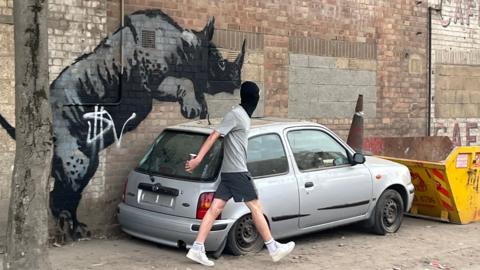 Man wearing a balaclava walks away after tagging the Banksy rhino picture with a white spray paint tag.