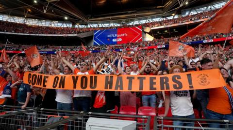Luton Town fans celebrating in the stands at Wembley Stadium after their side won promotion to the Premier League following the Sky Bet Championship play-off final in May 2023. They are holding a banner which reads: "Come on You Hatters". 