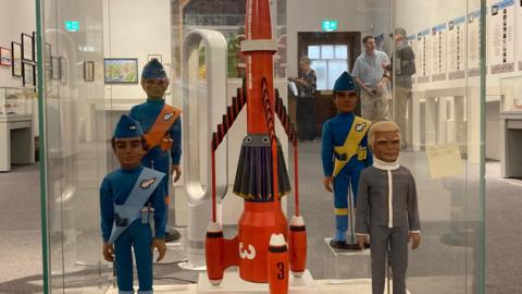 A large orange Thunderbirds rocket model displayed alongside Thunderbirds character figures at a museum exhibit.