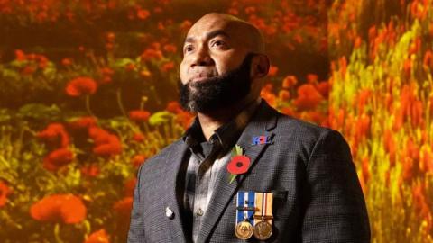 Nick Bennion, a former soldier wears a suit with his medals on the left lapel and a red poppy is stood in a room filled with poppy projections.
