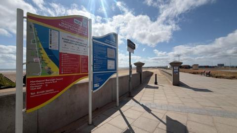 Signs at the promenade show a map of the beach and outline parking rules. The large sandy beach is sprawling in the background. 