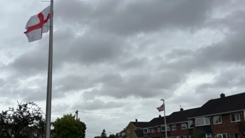 A St George's flag on a lamppost on the left-hand side of the road, and a union jack flag on a lamppost on the right-hand side of the road. There are houses on either side.