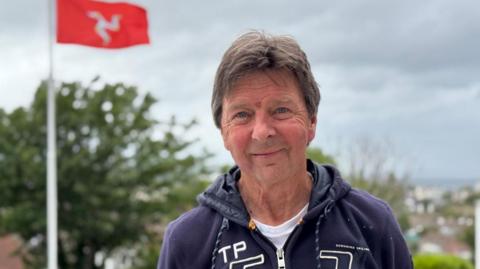 Glen Kinrade has short brown hair and is wearing a dark blue hoodie. He's standing outside with trees behind him.