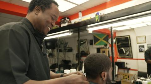 Ziggy cutting a customer's hair. He is smiling as he cuts hair, and Ziggy is wearing black top, with the customer sitting down.