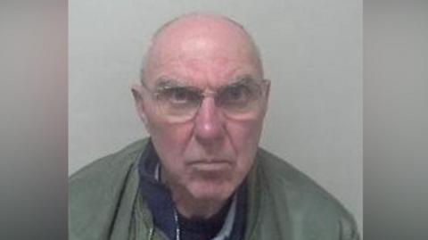 An elderly bald man in a mugshot. He is wearing a green jacket and glasses.