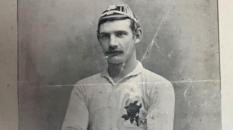A black-and-white picture of Little Billie Taylor wearing his England cap and England rugby shirt. The shirt has a large picture of a rose on the left-hand side.