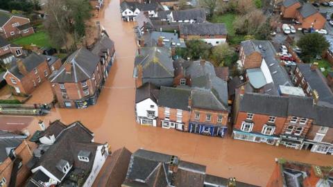 Flooded streets in Tenbury Wells. Long stretches of roads are under water, with water levels appearing several centimetres deep. The water is brown. There are no people or vehicles visible on the water. Shops, homes and businesses can be seen.