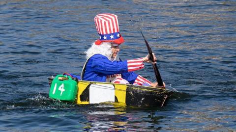 A man wearing fancy dress paddles a black and yellow home-made tin bath across a body of water.