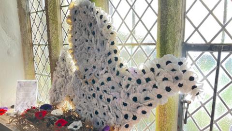 White poppies knitted together in the windowsill of the church. There is both purple, black and red poppies below it.