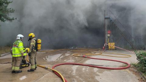 Fire fighters stand near a building which is almost completely hidden by thick black smoke.