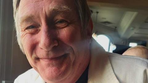 A close-up of a man in his 50s. He has a slightly red face and is smiling with his mouth closed. He has sideburns and a white collared shirt-jacket.