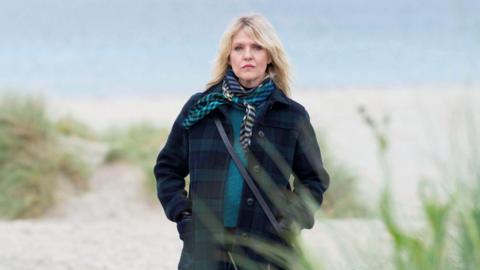 Ashley Jensen stands on a deserted Shetland beach, the sea grey behind her. She is wrapped up in a thick green and navy tartan wool coat with a teal woolly jumper and a patterned scarf. Her hands are in her pockets as she looks moodily at the camera.