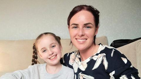 Selina Wallace has dark hair and is wearing a black top with white decorations. Her daughter Eliza has light brown hair in pigtails and wears a grey sweat shirt.