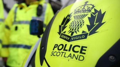 An image of 'POLICE SCOTLAND' lettering in bold, on a yellow police vehicle, with an officer dressed in yellow luminous uniform in the background.